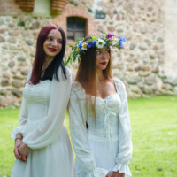 Two women in white dresses pose outdoors by a stone wall, one wearing a colorful flower crown and both smiling.