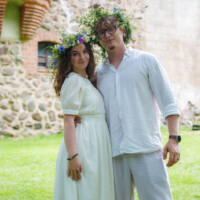 Couple wearing white outfits with flower crowns, smiling and embracing on a grassy lawn outdoors.