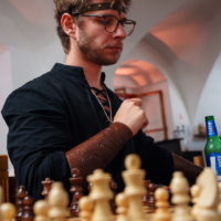 Man with glasses and a decorative headband sits at a chessboard, concentrating on the game in progress.