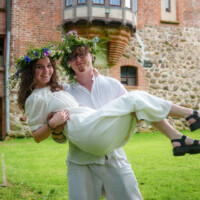 Couple in white outfits with flower crowns; man lifts smiling bride outdoors in front of a rustic brick building and grass.