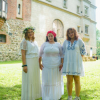 Three women in white dresses pose on a sunlit lawn, with a flower crown, red headscarf, and blue-embroidered outfit against a historic brick-and-stone building behind them.