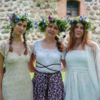 Three women wearing floral wreaths stand arm in arm on a lawn in front of a stone wall.