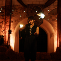 Male fire performer in black outfit spins flames from torches on a stage with warm lighting and arched background.