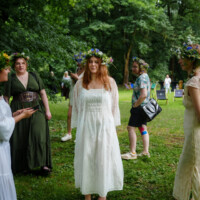 Young woman in a white dress with a flower crown stands barefoot on grass at an outdoor gathering in a park.
