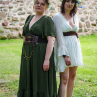 Two women in medieval-style dresses pose outdoors in front of a stone wall, wearing flower crowns and braided belts.