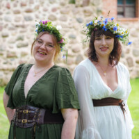 Two women in flower crowns pose outdoors in front of a stone wall, smiling; one wears a green dress with a corset belt, the other a white dress with a brown sash.