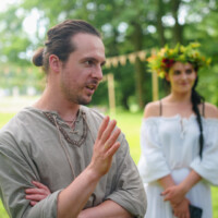 Man in a loose gray tunic gestures while speaking outdoors, with a woman in a white dress and flower crown standing in the background.