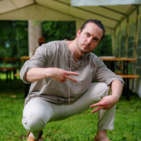 Man crouching on grass under a white event tent, making peace signs with both hands, wearing a beige tunic and light pants.