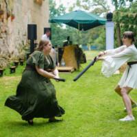 Two women in period costumes duel with foam swords on a grassy lawn, near a stone wall and festive bunting.