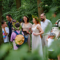 Group of people in a forest gathering, several wearing flower crowns and light, airy clothing, smiling and chatting.