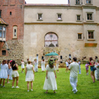 Group of guests in white dresses standing in a circle on a grassy lawn in front of a historic courtyard building.