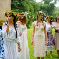 Women wearing flower crowns stand outdoors; one speaks into a microphone during a celebration.