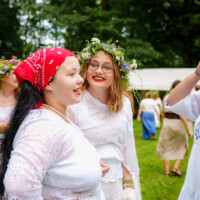 Women in white lace dresses wearing floral crowns laugh and chat at a sunny outdoor festival in a park.
