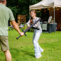 Young woman playfully swings a foam sword at a man during an outdoor festival, with a white tent and brick wall in the background.