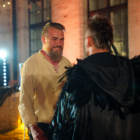 Two men at a festive indoor event smile at each other; one in a beige lace-up shirt, the other in a black feathered cape.