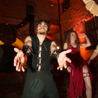 Two costumed individuals dance toward the camera at night in a courtyard lit by string lights, a man in a black buckle vest and wide pants and a woman in a red dress with a slit.