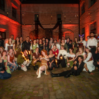 Large group in medieval costumes posing in a cobblestone courtyard at night with string lights and red uplighting; a central performer holds torches links the scene together.