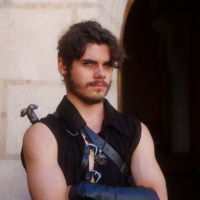 Young man cosplaying as a medieval knight, arms crossed, standing near an arched stone doorway with a sword hilt visible over his shoulder.