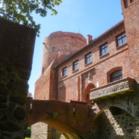 Red brick castle with a round tower connected by a stone-arched bridge over a green lawn under a blue sky.