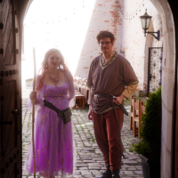 Two people in medieval costumes stand in a stone archway on a cobblestone street, with string lights and tables in the background.