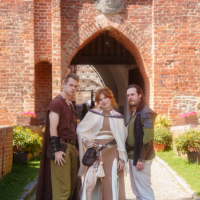 Three people in period costumes pose at a brick archway, medieval outfits with capes and armor stands on a cobblestone path.