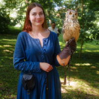 Woman in a blue dress stands in a sunny park with an owl perched on her gloved left arm, looking at the camera.