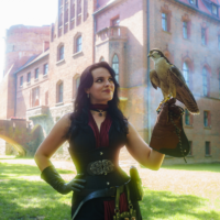 Woman in a black and red fantasy costume, wearing leather gloves, poses outdoors with a falcon perched on her gloved hand before a brick castle-like building.