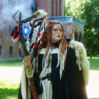 Cosplay performer in a black-and-white gothic gown with fur cloak, holding a tall staff wrapped in twine and feathers, glowing blue orb at the top outdoors near a brick castle.