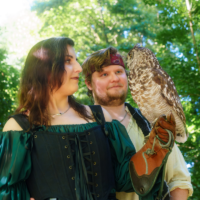 Two costumed adults stand in a sunlit forest, a woman in a green and black medieval dress beside a man with an owl perched on his gloved arm.