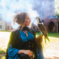 Woman in a blue and black medieval-style outfit holding a falcon on her gloved hand, smoke filling the air behind a castle-like brick building.