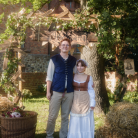 Two people in medieval costumes stand under a leafy wooden arch, with hay bales nearby in a sunny yard.