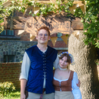 Couple in Renaissance costumes posing under an ivy-covered wooden arch with a carved sign, outdoors.