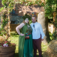 Couple in medieval-inspired clothing posing under a ivy-covered wooden arch at an outdoor festival, with a wicker basket and hay at their feet.