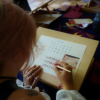 Person writing runic letters on a practice sheet at a wooden desk in a studio setting.
