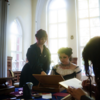 Two women in vintage dresses study documents at a wooden desk in a bright room with tall arched windows and a chandelier.