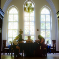 Four people sit around a round table reading menus in a bright, elegant room with tall arched windows and a grand chandelier above.