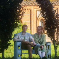 Two friends sit on a wooden bench under a leafy arch in a sunny garden, each holding a beer, with a brick building visible in the background.