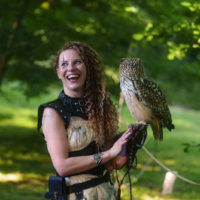 Woman in a medieval-style costume smiles while an owl perches on her gloved arm outdoors in a parkfest setting.