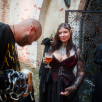 Woman in a black lace gown with a tiara, holding a beer and showing colorful tattoos at a brick courtyard event.