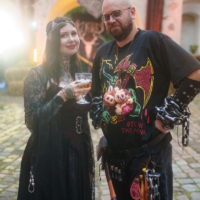 Two costumed adults posing outdoors, the woman in a black lace gown with a jeweled headpiece holding a glass of wine, the man in a black shirt with chains and prop weapons.