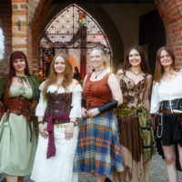 Five women in medieval costumes posing together outdoors in front of a brick archway and iron gate, smiling for the camera.