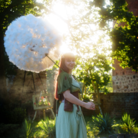 Woman in a green vintage dress outdoors, holding a giant white dandelion-like puffball against a sunlit background.