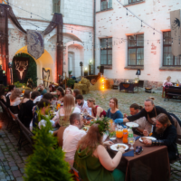Group of people dining at a long table in a cobblestone courtyard, string lights overhead, old brick building in background.