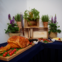 Buffet spread with a roasted meat on a cutting board, cherry tomatoes, herbs, and a carving knife, against a backdrop of potted plants and wooden crates.