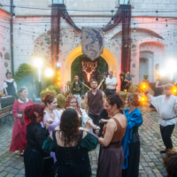 People in colorful medieval costumes dance in a cobblestone courtyard forming a circle during a festival, with string lights and a bannered arch in the background and warm torches illuminating the scene.