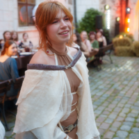 Young woman in a medieval-inspired beige and brown outfit smiling at an outdoor event with blurred crowd in the background.