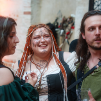 Three people in cosplay at a party; center person with orange braided hair and face paint smiles while others chat.|