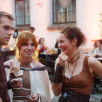 Three adults in period-costume outfits chat and laugh at an outdoor festival, with a brick building in the background.