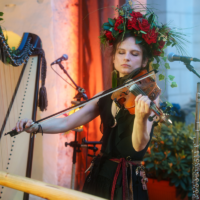 Female violinist wearing a vibrant red flower crown, bowing the violin with eyes closed, on a stage setup with microphones in the background.