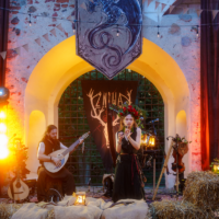 Woman with a flower crown sings into a microphone on a rustic stage, guitarist beside her; hay bales in the foreground under string lights and a stone arch.